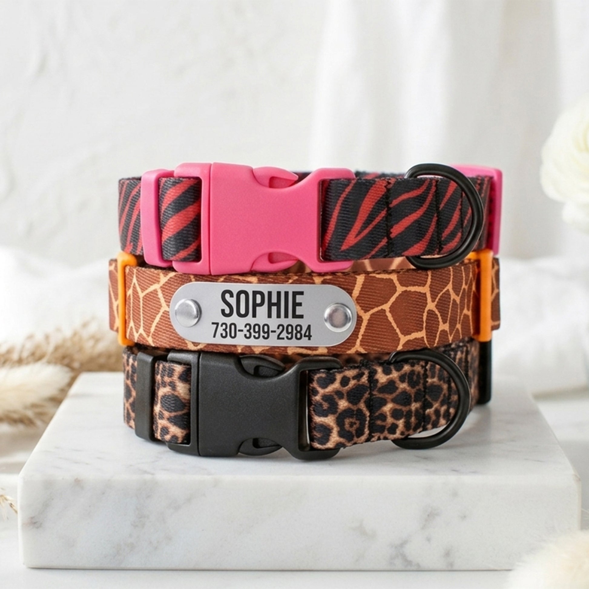 Three animal print dog collars with a pink buckle and a nameplate on a marble surface.