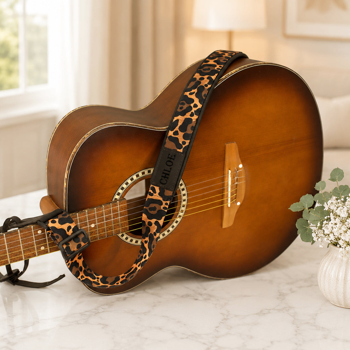 Acoustic guitar with a leopard print strap on a marble surface, with a blurred indoor background.