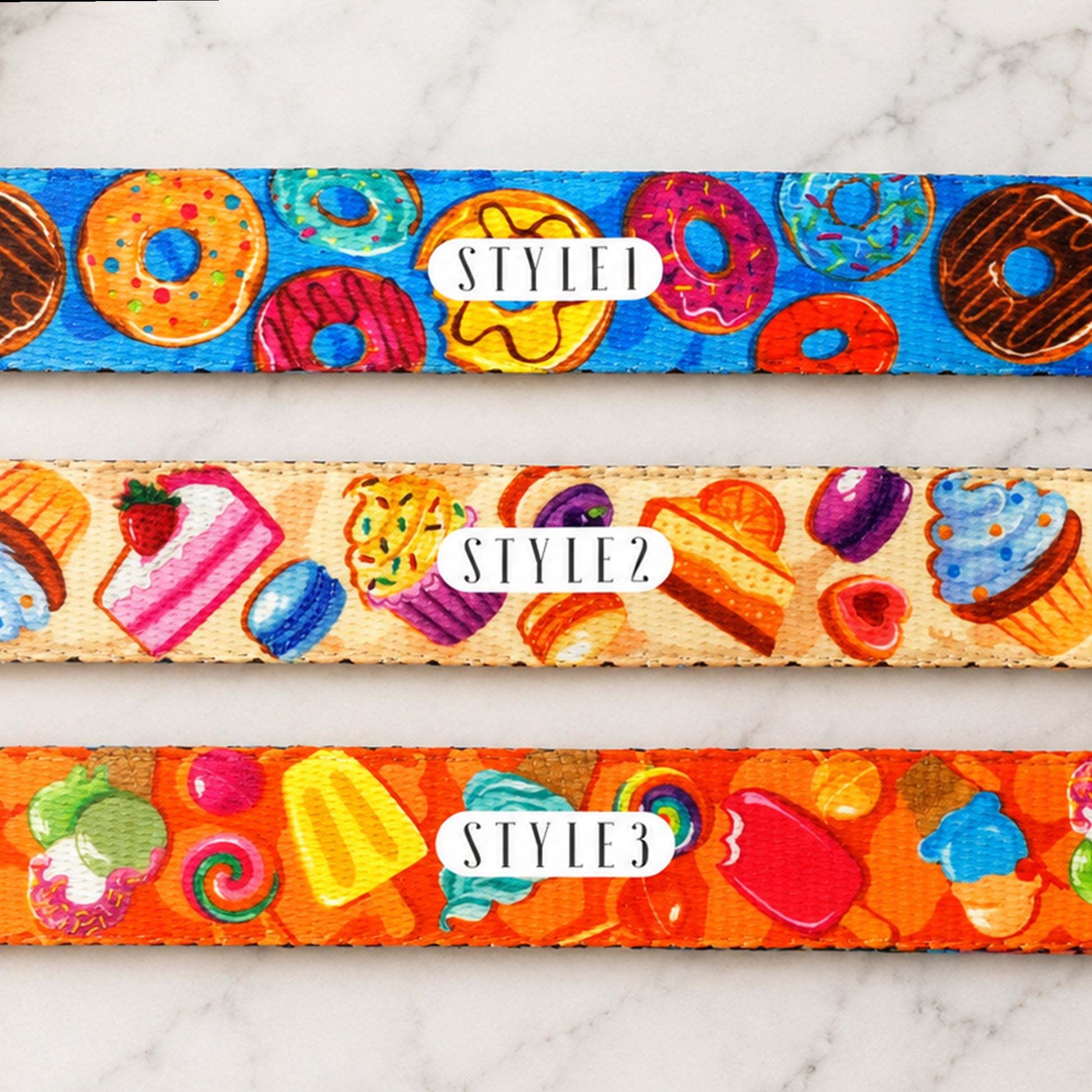Three colorful belt straps with donut, cake, and candy patterns on a marble surface.