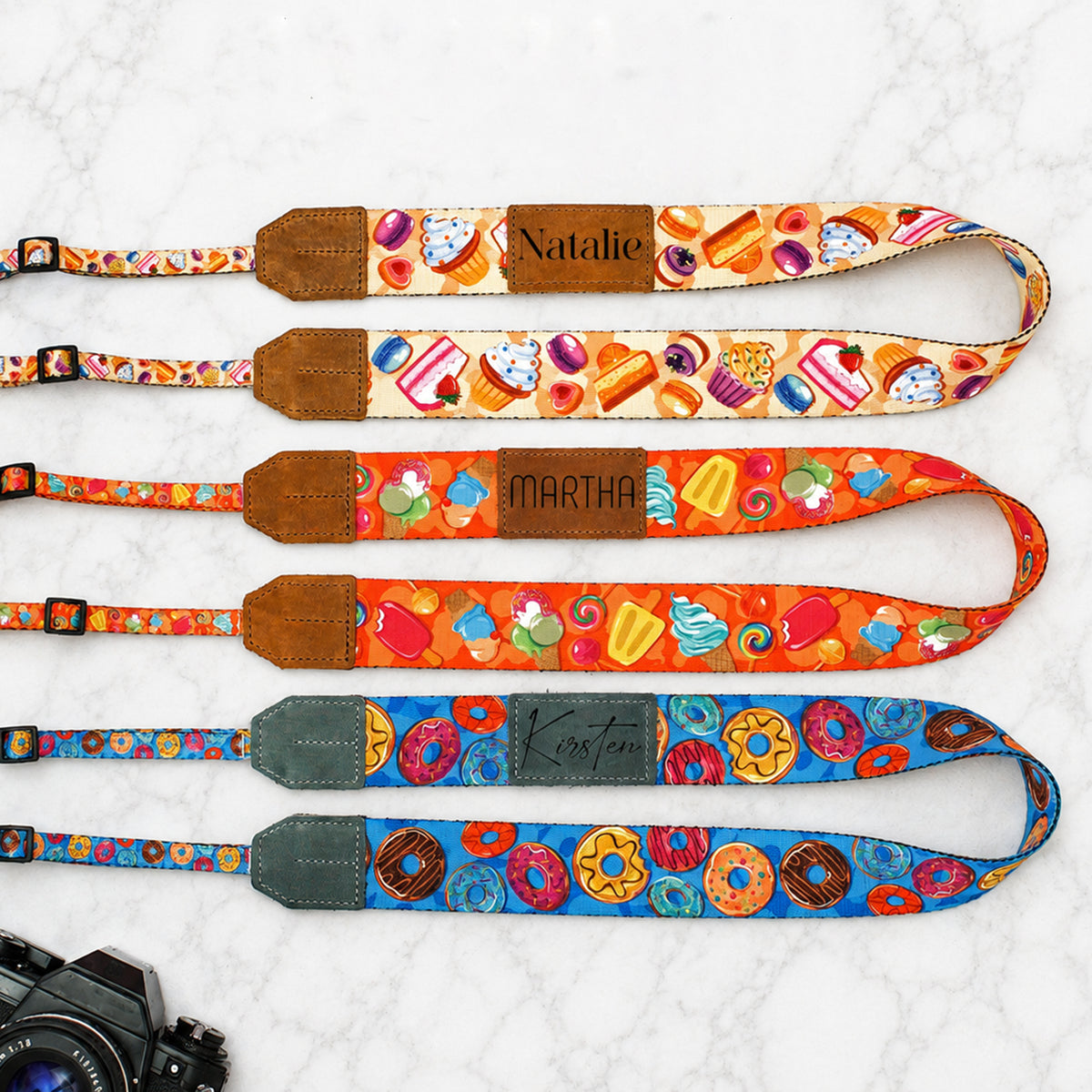 Four colorful camera straps with different patterns and names on a textured white background.