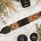 Camera strap with decorative pattern and Nikon logo, placed on a marble surface with camera lenses and leaves.