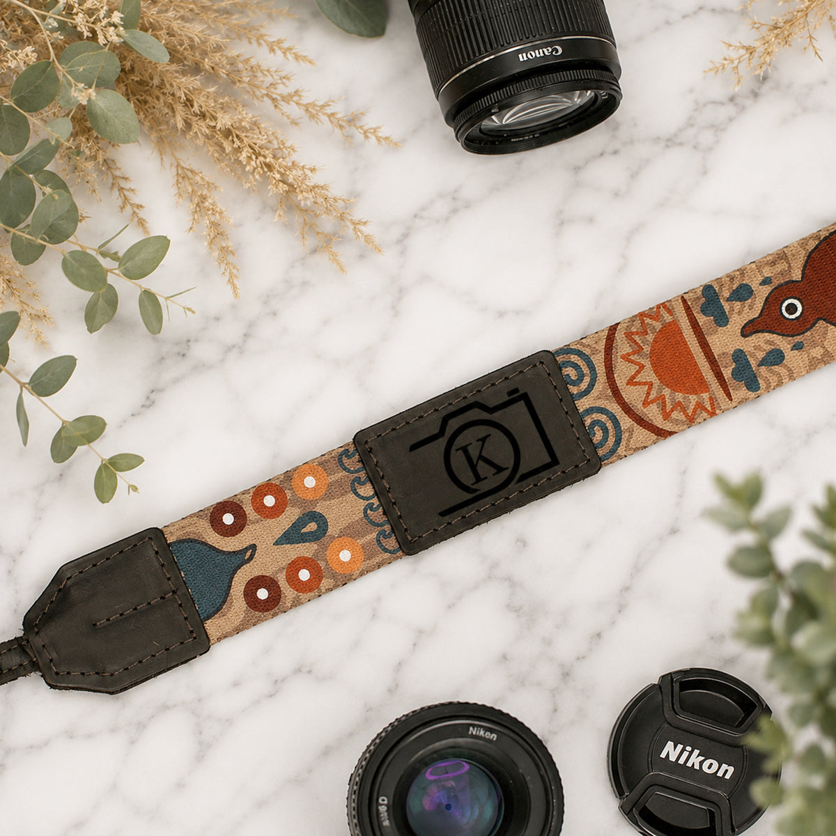 Camera strap with decorative pattern and Nikon logo, placed on a marble surface with camera lenses and leaves.