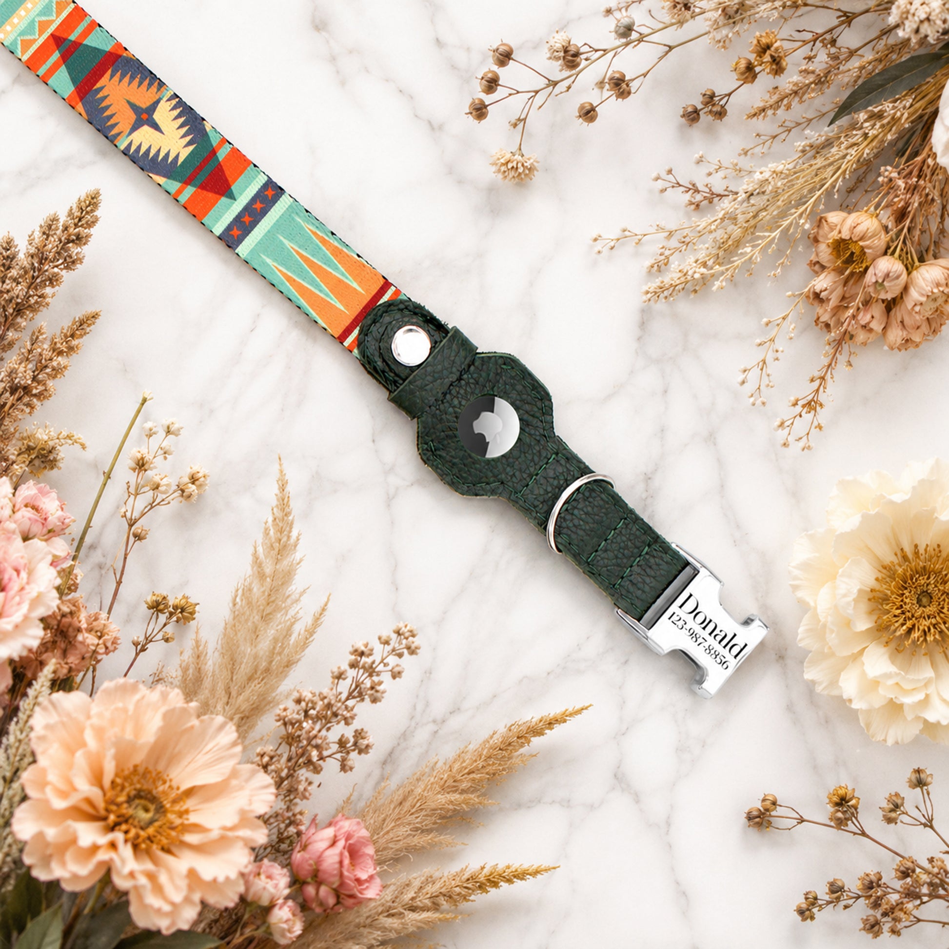Dog collar with colorful pattern and engraved name tag on a marble surface with flowers and dried plants.