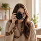 Woman holding a camera with a leopard print strap in a cozy indoor setting