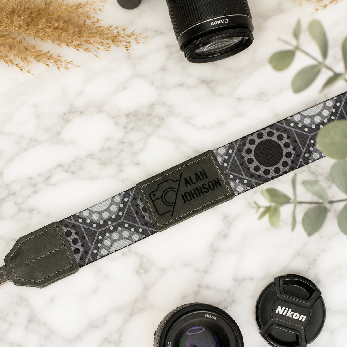 Camera strap with personalized engraving on a marble surface with camera lenses and leaves.