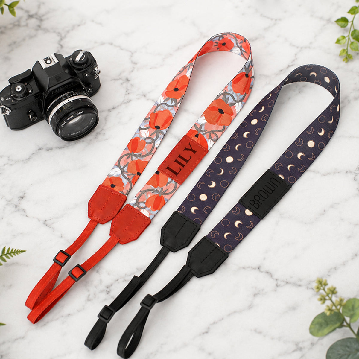 Two camera straps with floral and moon designs on a marble surface with a camera.