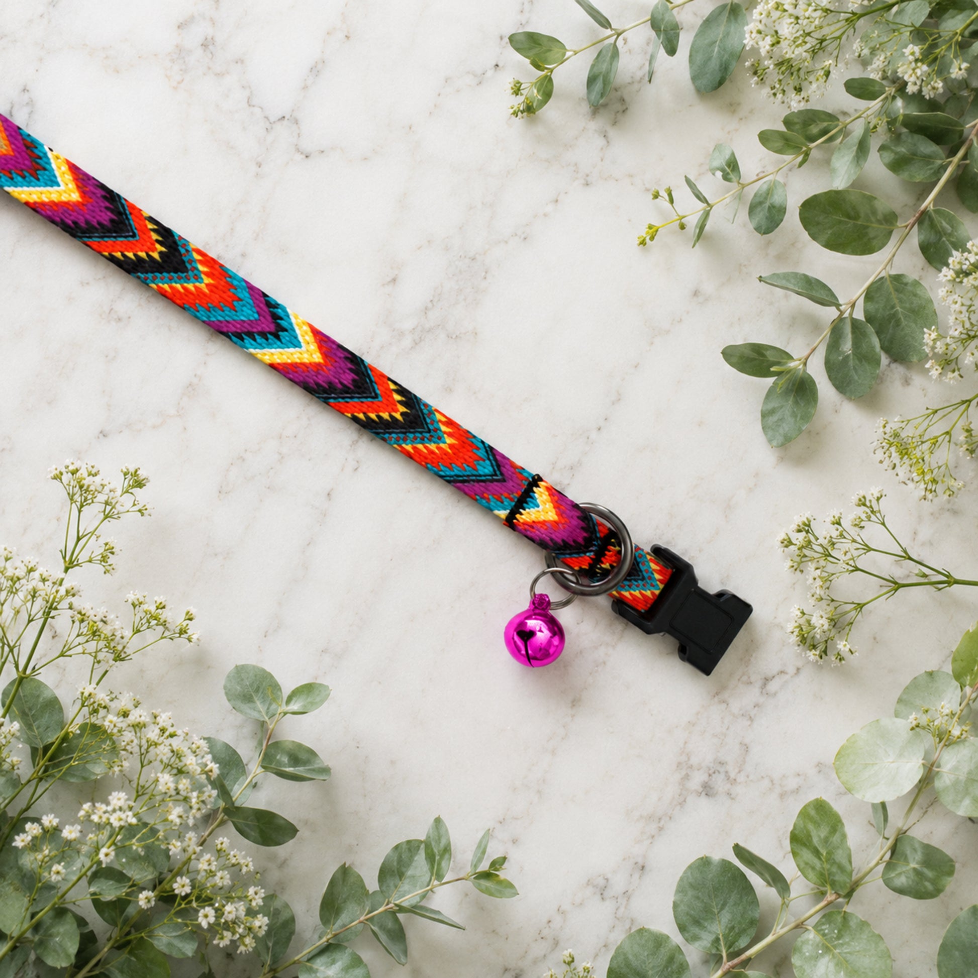 Colorful braided dog collar with a bell on a marble surface surrounded by greenery