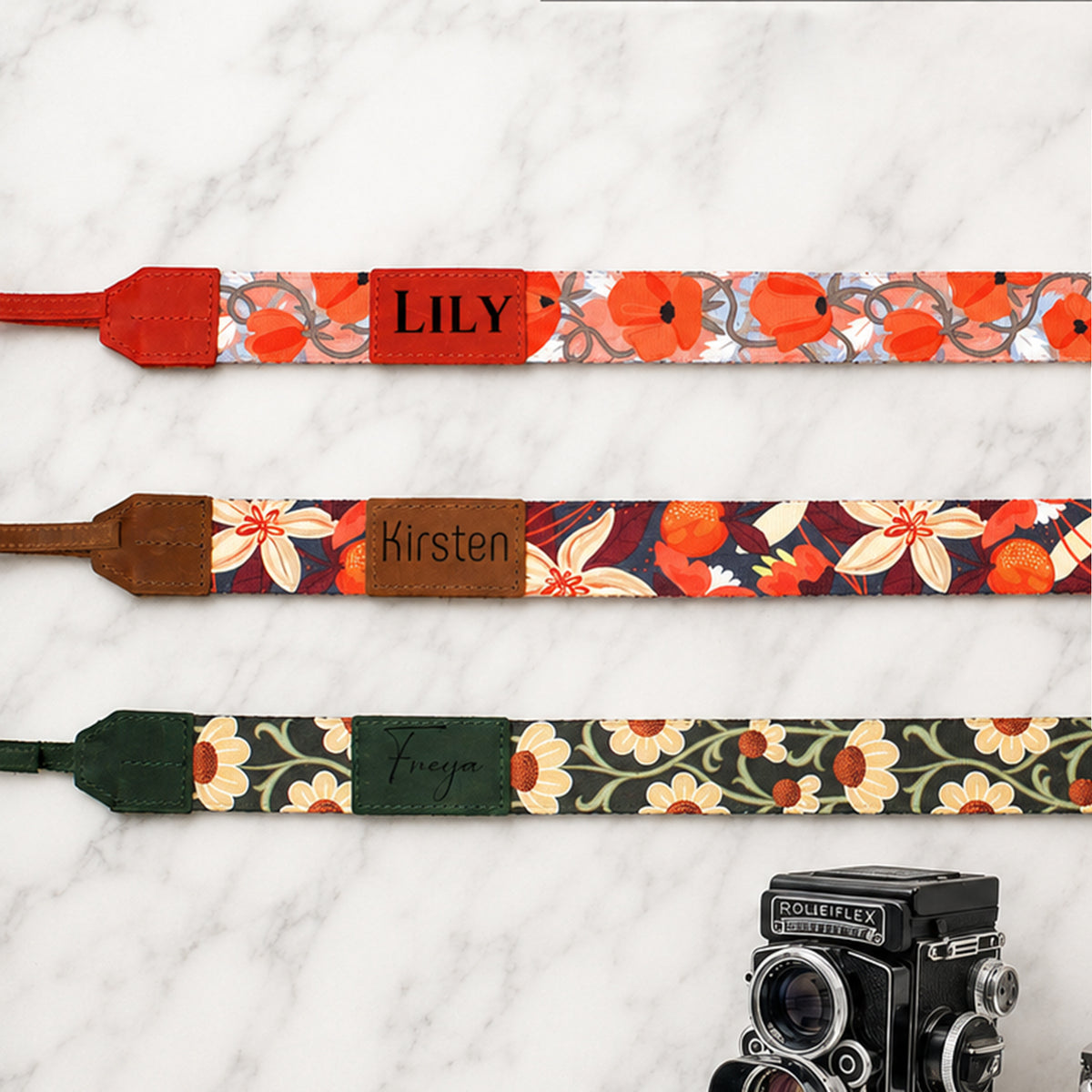 Three floral camera straps with names engraved on them, placed on a white surface with an old-fashioned camera.