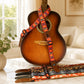 Acoustic guitar with a colorful strap on a light surface, with a window and plant in the background.