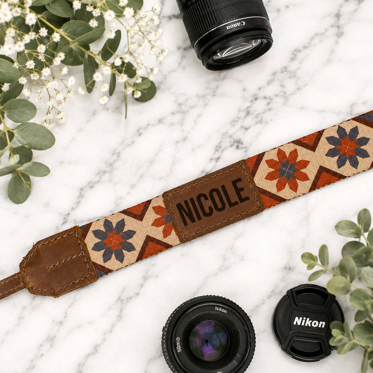 Camera strap with 'NICOLE' engraving, surrounded by camera lenses and greenery on a marble surface