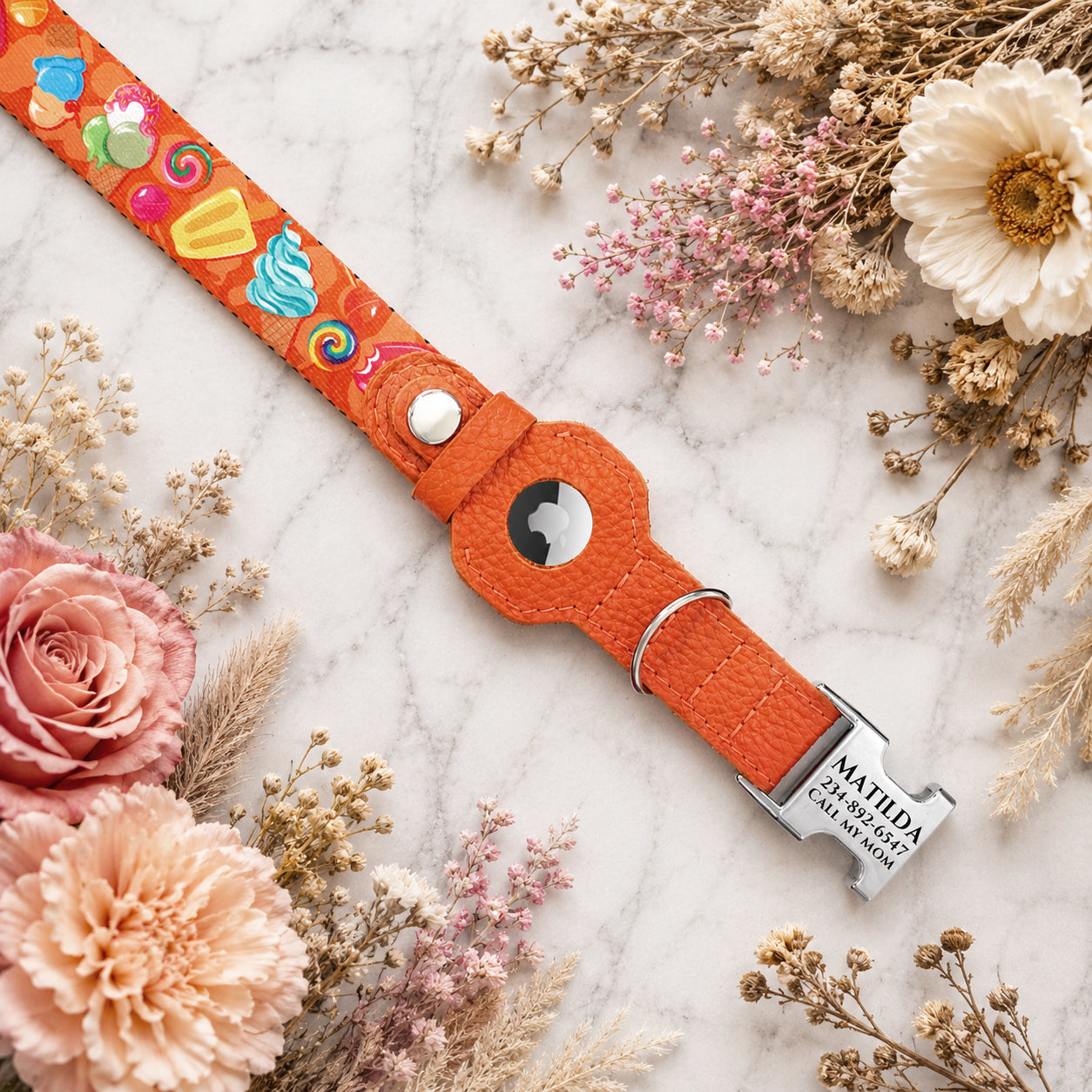 Orange dog collar with colorful designs and a silver tag on a marble surface with flowers.