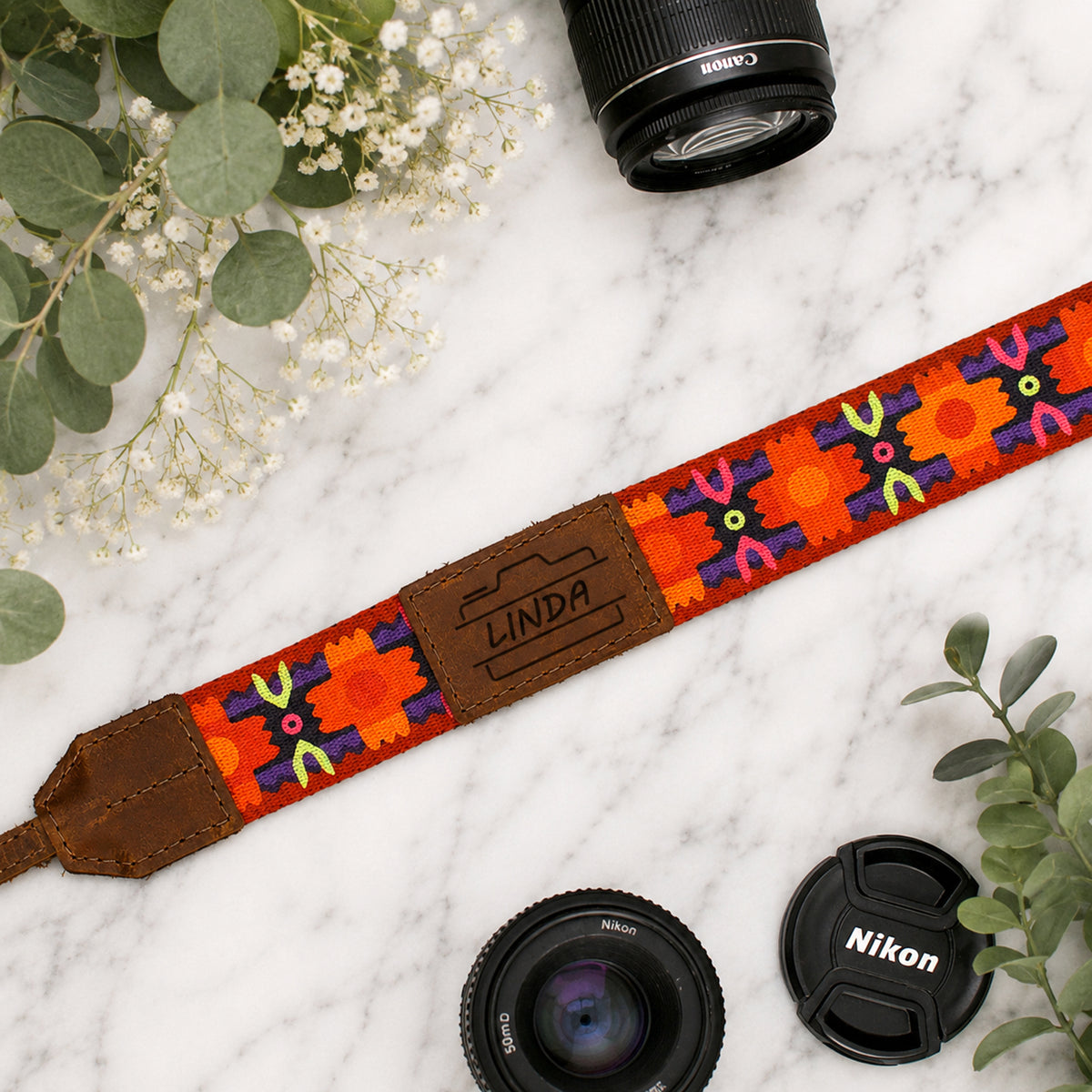 Colorful camera strap with 'Linda' branding on a marble surface with camera lenses and leaves.