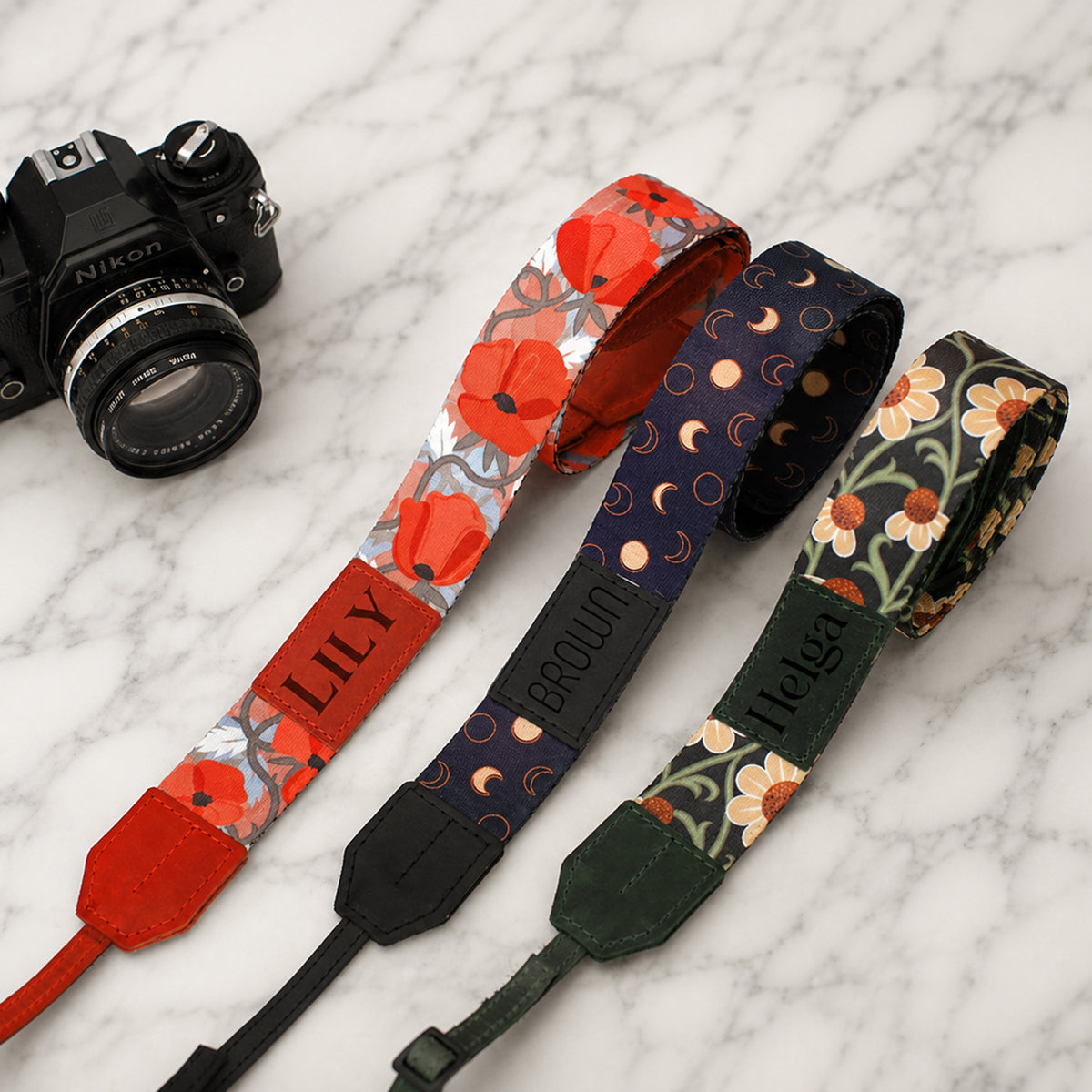 Three floral-patterned camera straps with 'Lily', 'Brown', and 'Mila' branding on a marble surface with a Nikon camera.