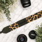 Leopard print camera strap with 'Daniela' engraving, surrounded by camera lenses and leaves on a marble surface.