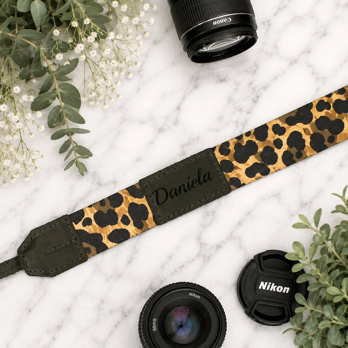 Leopard print camera strap with 'Daniela' engraving, surrounded by camera lenses and leaves on a marble surface.