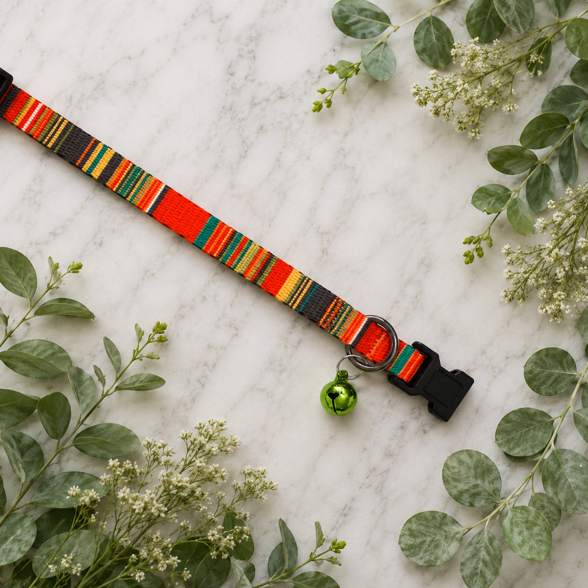 Colorful striped dog collar with a green bell on a marble surface with green leaves.