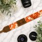Camera strap with floral pattern and leather accent on a marble surface with camera lenses and flowers.