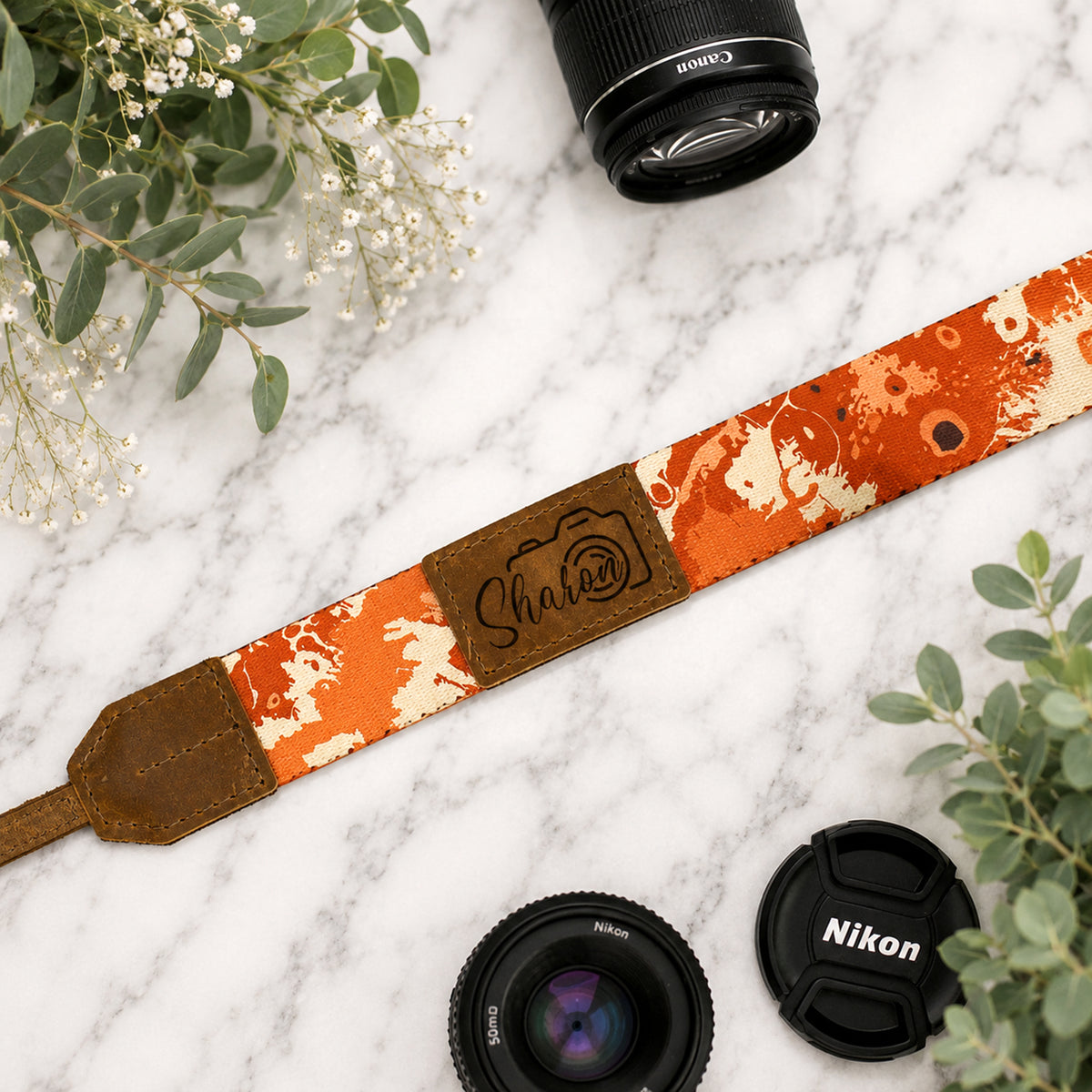 Camera strap with floral pattern and leather accent on a marble surface with camera lenses and flowers.