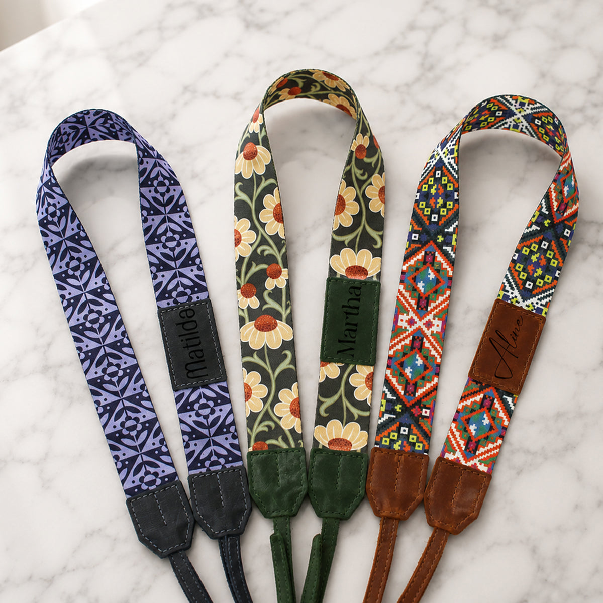 Three colorful camera straps with floral and geometric patterns on a marble surface.