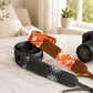 Camera strap with floral pattern on a marble surface, with a camera and lens in the background.