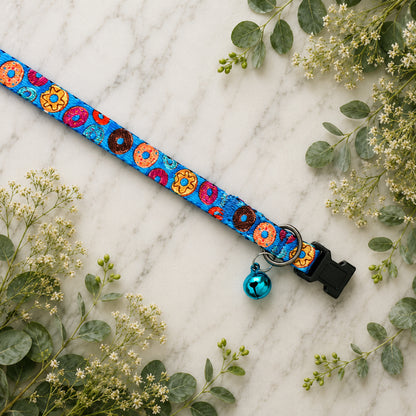 Colorful donut-patterned dog collar with a bell on a marble surface with greenery.
